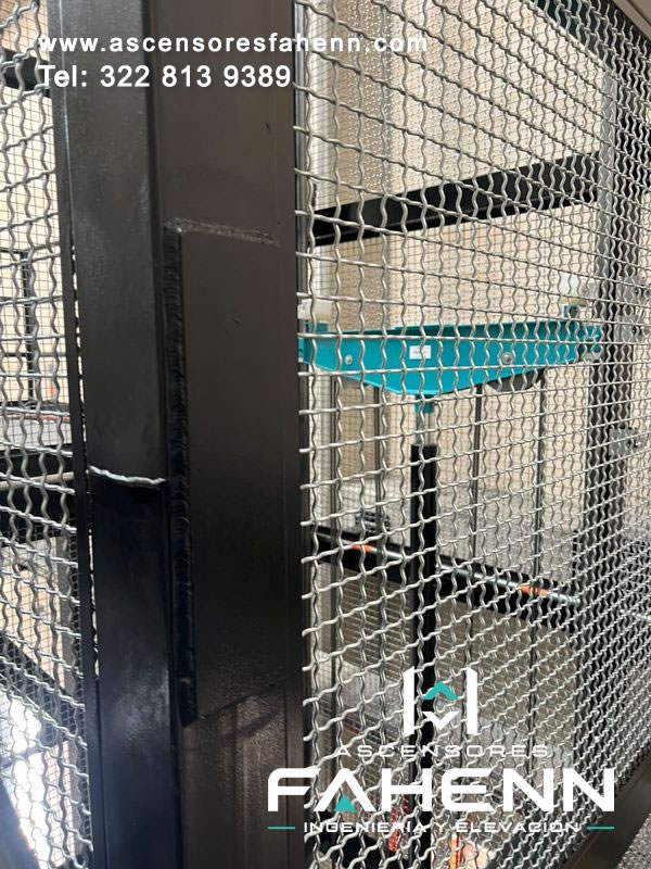 Sistema hidráulico de ascensor industrial con componentes metálicos visibles detrás de una malla protectora.