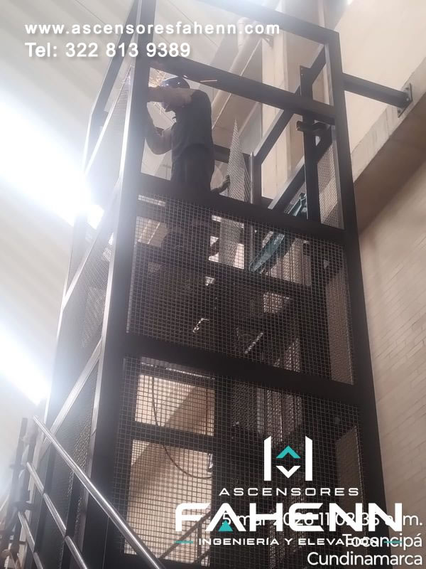 Montaje técnico de elevador industrial con operario trabajando en la parte superior de una estructura metálica.