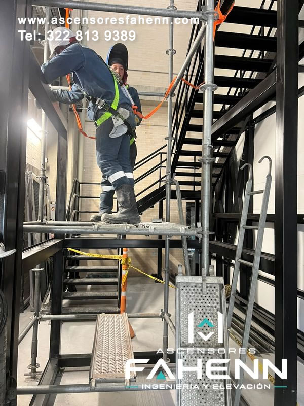 Técnicos con arneses y líneas de seguridad trabajando sobre un andamio durante el montaje de un ascensor industrial.