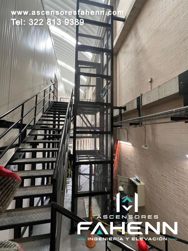 Elevador industrial vertical con estructura negra de malla instalado junto a una escalera metálica.