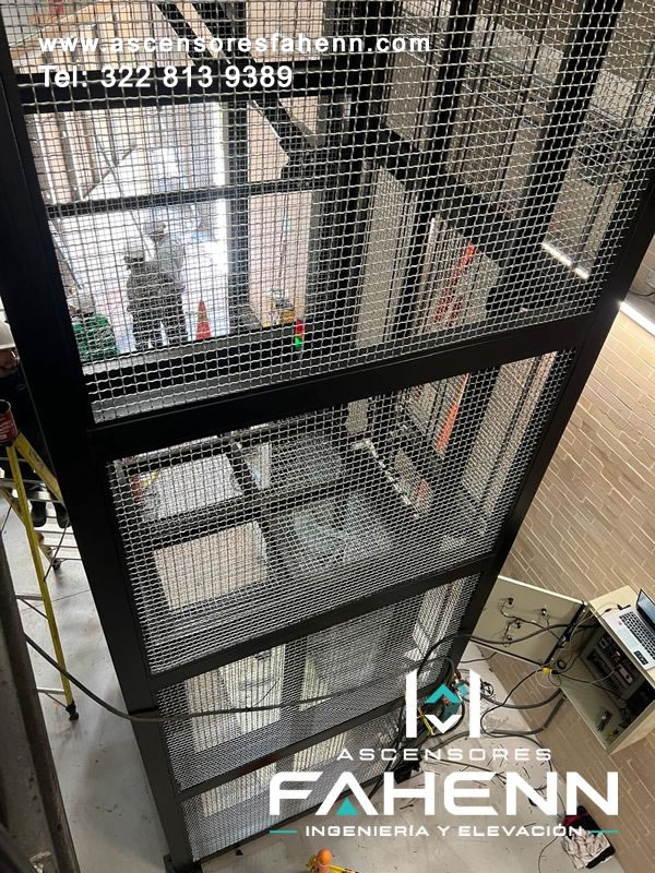 Estructura protectora de ascensor de carga con perfiles negros y paneles verticales de malla metálica reforzada.
