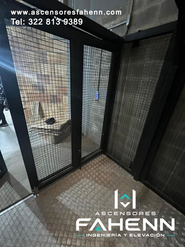 Ascensor para mercancías con cabina de malla, perfiles negros, puerta abierta y piso metálico antideslizante.