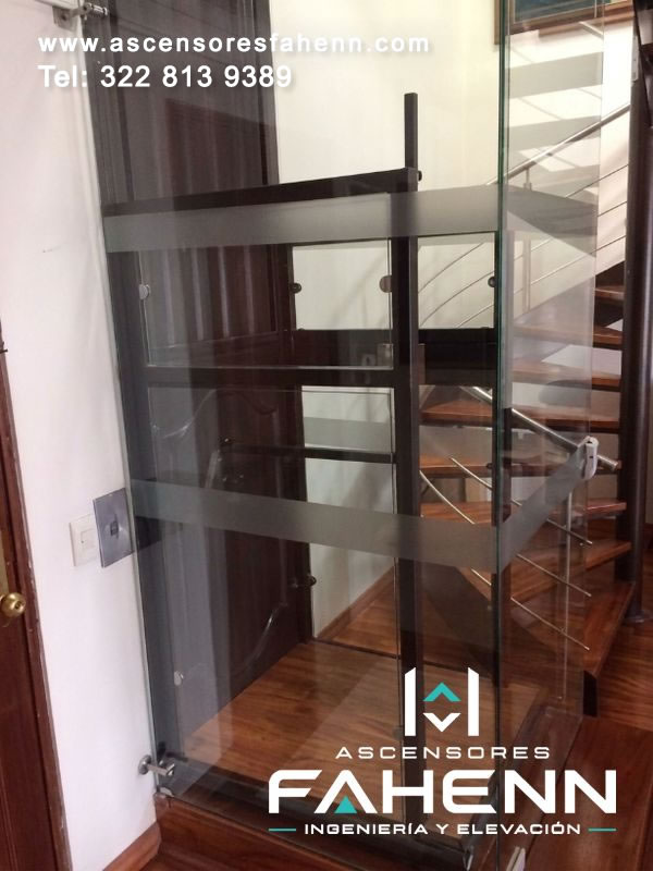 Ascensor para casa con estructura de vidrio, plataforma compacta tipo madera e instalación junto a escaleras.