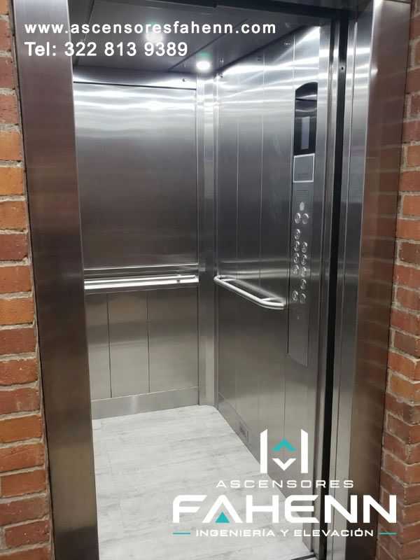 Cabina para ascensor con revestimiento de acero inoxidable, dos pasamanos laterales, panel de control multinivel y acceso frontal despejado.
