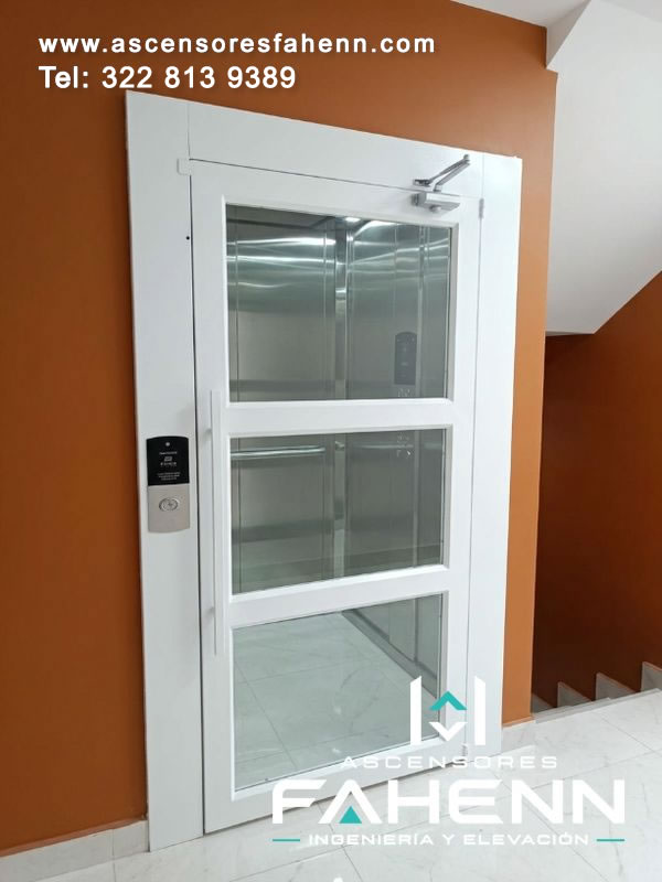 Elevador residencial con puerta acristalada de marco blanco, tres secciones transparentes, cierre superior y cabina metálica visible desde el descanso.