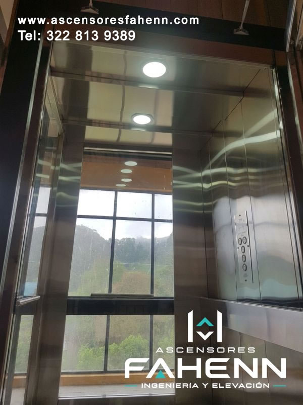 Interior de cabina panorámica con vidrio de piso a techo, paisaje exterior, revestimiento de acero inoxidable y luces redondas empotradas.
