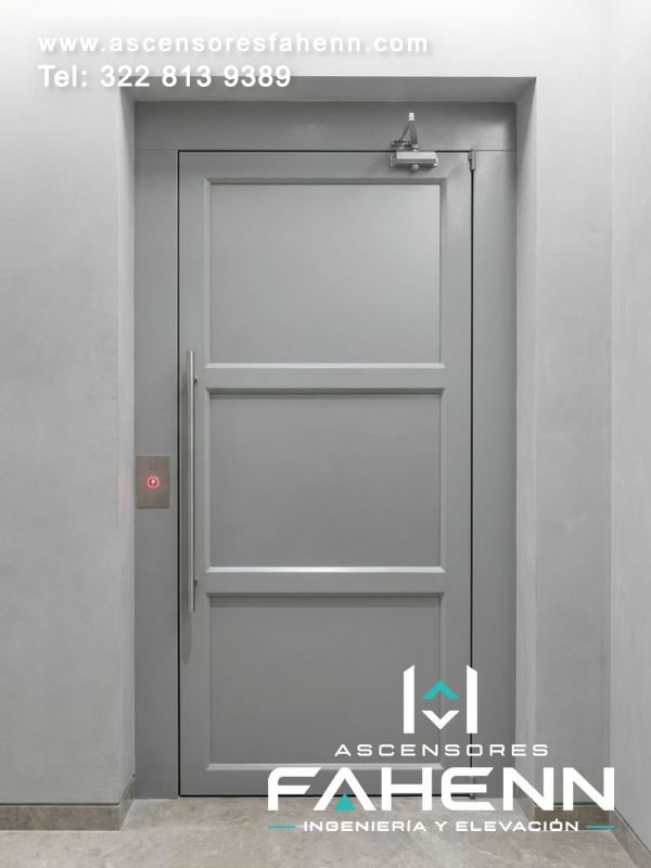 Puerta para ascensor con marco gris, hoja de diseño sólido, manija alargada, mecanismo superior de cierre y pulsador LED encendido junto al acceso.