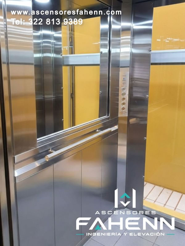 Cabina de ascensor con espejo, controles circulares alineados, baranda horizontal y revestimiento de acero combinado con acentos de color amarillo.