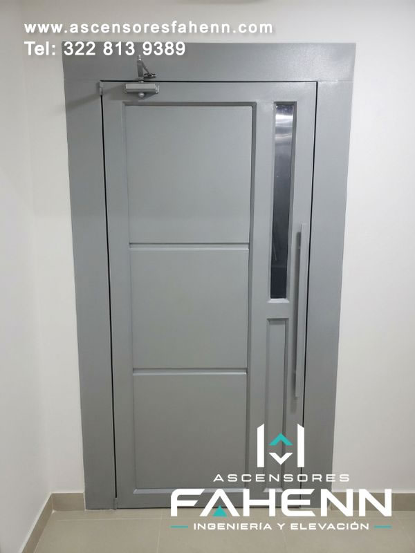 Puerta para ascensor con hoja metálica, mirilla vertical, manija lateral y cierrapuertas instalado en la parte alta de un marco compacto.