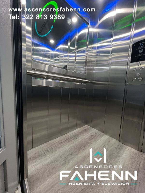 Cabina de ascensor con acero inoxidable reflectivo, espejo lateral, baranda metálica, piso tipo madera y luz LED de varios colores en el techo.