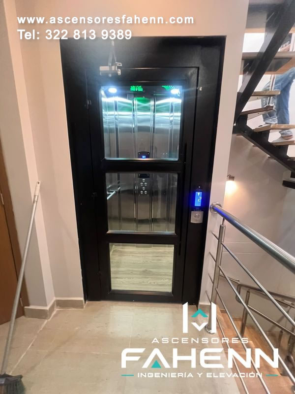 Elevador residencial junto a una escalera con puerta de tres paños de vidrio, interior reflectivo iluminado y pantalla exterior en el piso 2.