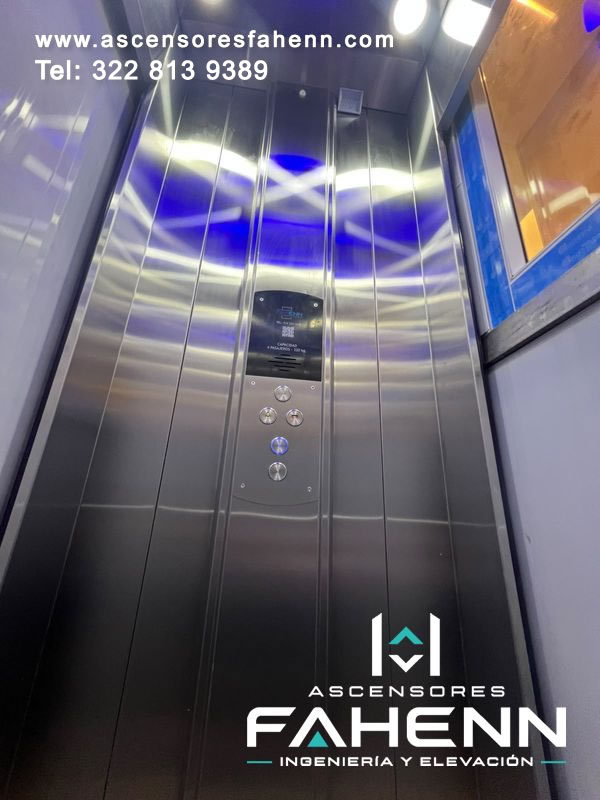 Cabina de ascensor para cuatro personas con revestimiento de acero, pulsadores circulares, reflejos LED azules y ventana ubicada en un costado.