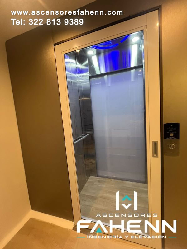 Puerta transparente para ascensor con marco metálico claro y visual hacia un interior de acero, iluminación azul y superficie blanca decorativa.