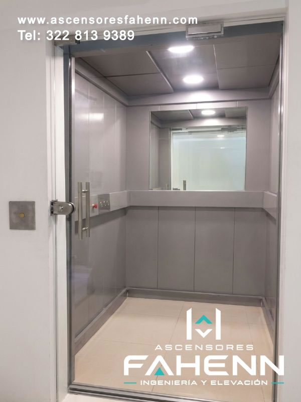 Cabina de ascensor con espejo, revestimientos grises, puerta transparente, luces circulares y controles integrados sobre una franja horizontal.