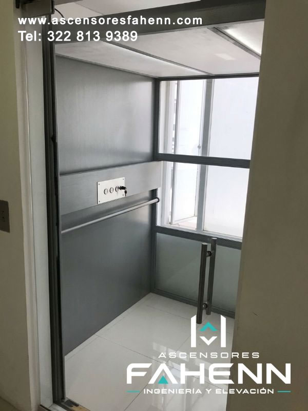 Cabina transparente para elevador de vivienda con baranda lateral, mandos compactos, piso claro y grandes superficies de vidrio hacia el exterior.