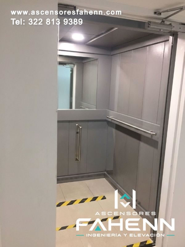Interior amplio de ascensor con espejo, revestimientos grises, baranda metálica, puerta abierta y franjas de señalización sobre el piso.