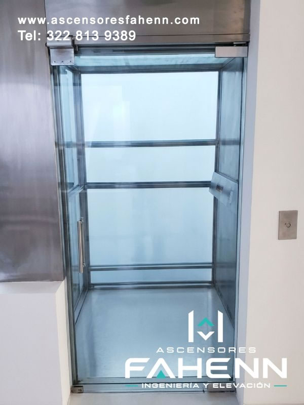 Cabina acristalada de ascensor con amplios paños transparentes, perfiles metálicos, tirador vertical y acceso despejado desde un corredor blanco.