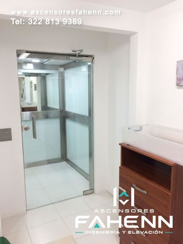 Ascensor para interiores con puerta transparente, marco metálico, cabina clara y superficies acristaladas integradas junto al mobiliario residencial.