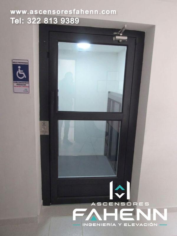Puerta acristalada para plataforma de movilidad reducida con marco negro, llamada exterior y visual hacia un corredor interior de tonos claros.