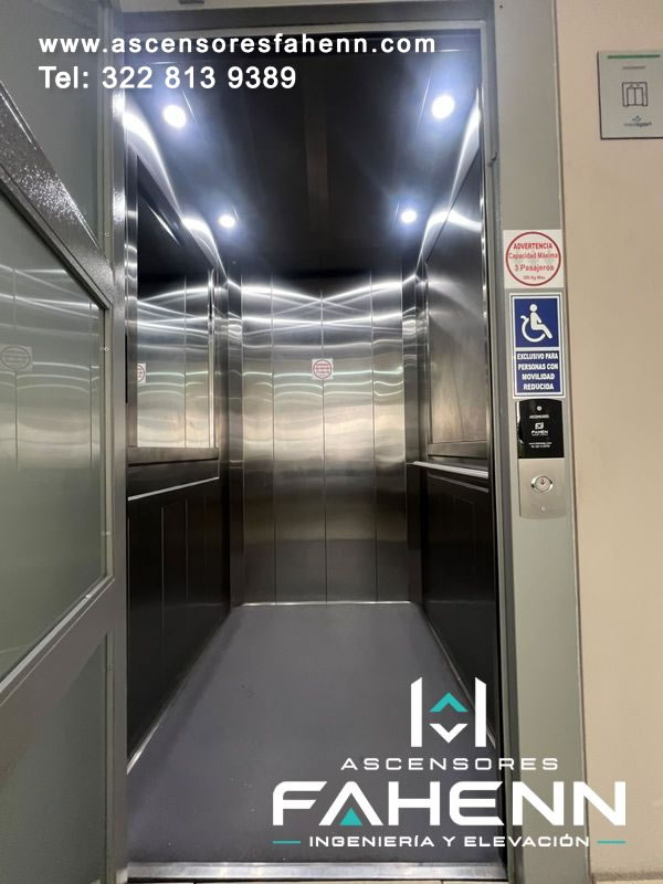 Cabina accesible de ascensor en acero reflectivo con piso gris, luces superiores, barandas laterales y señal para personas con movilidad reducida y acceso amplio.