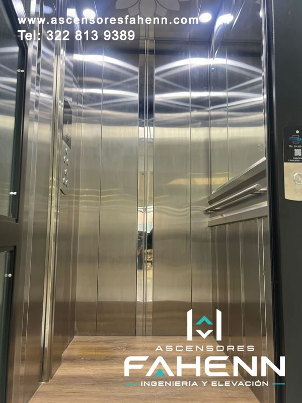 Cabina de ascensor en acero brillante con paneles reflectivos, botonera lateral, pasamanos, luces circulares y piso con acabado tipo madera y acabado premium.