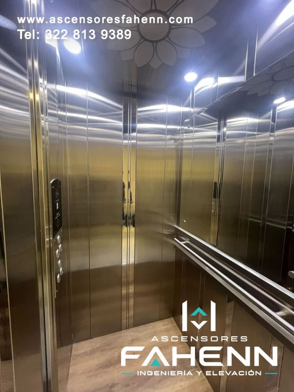 Interior de ascensor moderno con espejo, pasamanos doble, paneles de acero reflectivo, botonera vertical, techo floral y piso tipo madera de acabado premium.