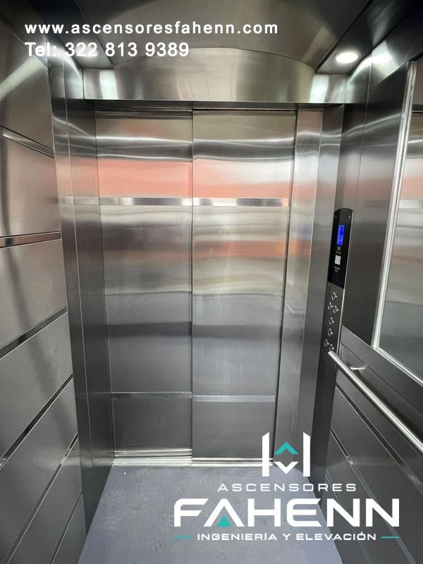 Cabina de elevador en acero inoxidable con puertas automáticas cerradas, pantalla azul, botonera vertical, espejo y pasamanos lateral de acabado moderno.