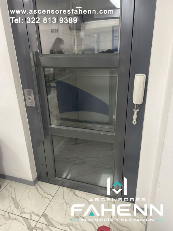 Cerramiento acristalado para ascensor con estructura metálica gris, estación de llamada marcada con 3 y equipo telefónico de comunicación lateral para atención cercana.