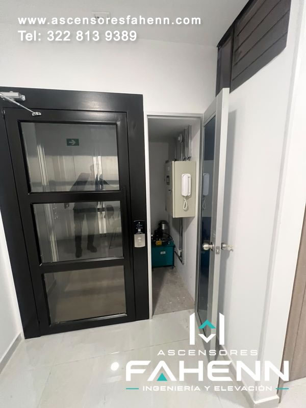 Ascensor residencial compacto con puerta negra de tres vidrios, llamada exterior y cuarto técnico abierto con unidad hidráulica y gabinetes en un espacio compacto.