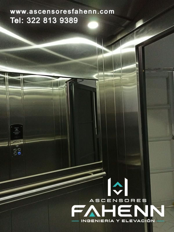 Interior de ascensor con gran espejo, pasamanos doble de acero, paneles metálicos oscuros y reflejos luminosos producidos por la luz del techo.
