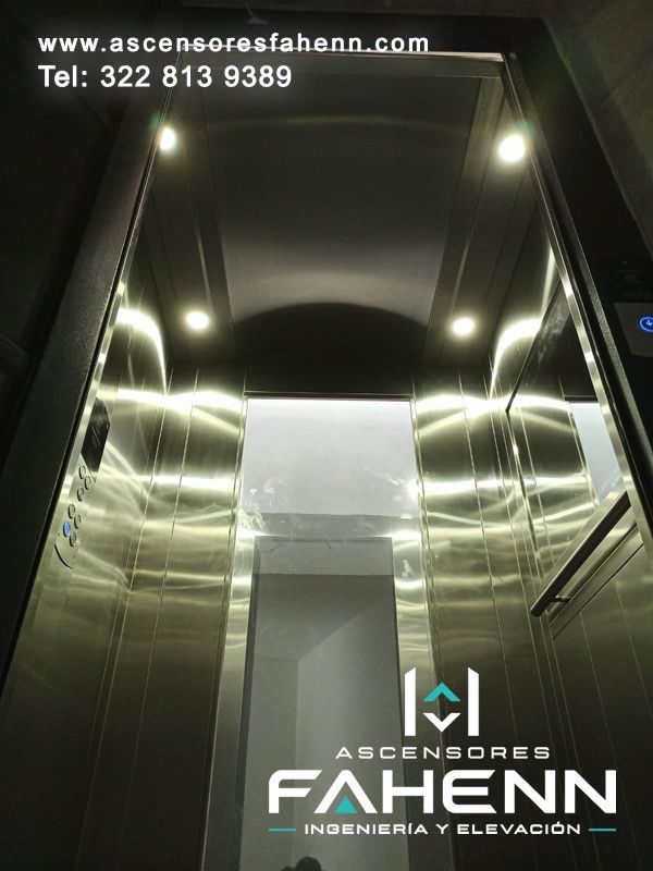 Cabina premium de acero vista desde abajo, con paneles reflectivos, ventana panorámica vertical, botonera lateral, pasamanos y luces LED superiores.