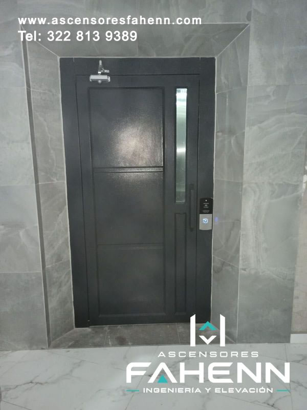 Puerta negra de ascensor residencial con visor vertical estrecho, manija recta, cierrapuertas superior y pulsador iluminado en un muro de placas grises.