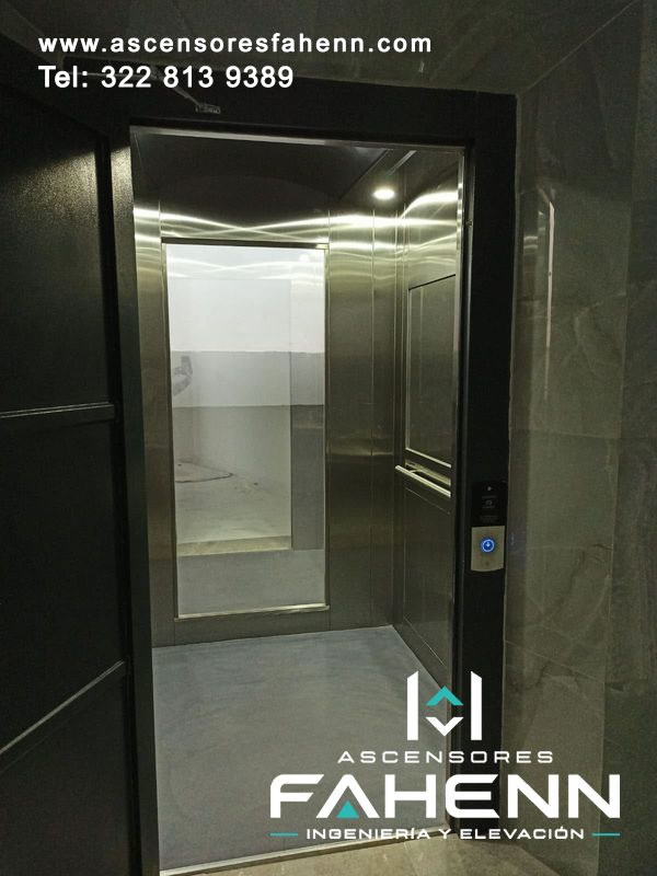 Ascensor residencial abierto con cabina de acero inoxidable, espejo posterior de gran formato, pasamanos, luces en el techo y piso gris continuo.