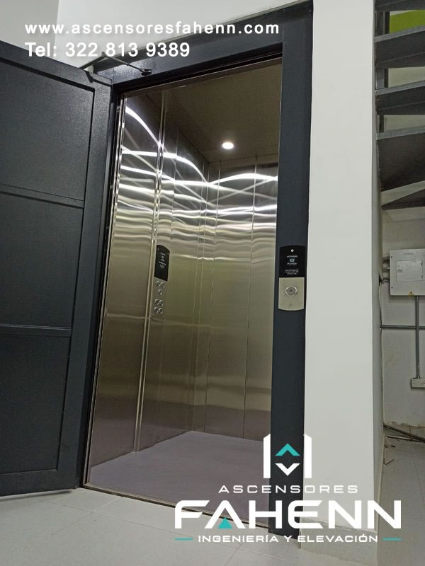 Cabina premium abierta con paneles de acero inoxidable, luces LED reflejadas, botonera lateral, piso gris y marco exterior de color negro.