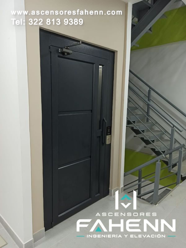 Puerta negra de ascensor con visor vertical estrecho, paneles rectangulares, manija, control lateral y mecanismo de cierre instalado arriba.
