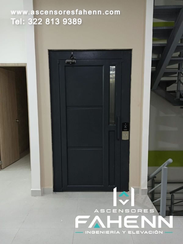 Puerta residencial negra de ascensor con visor vertical de vidrio, paneles geométricos, cierre superior, manija y pulsador exterior de llamada.