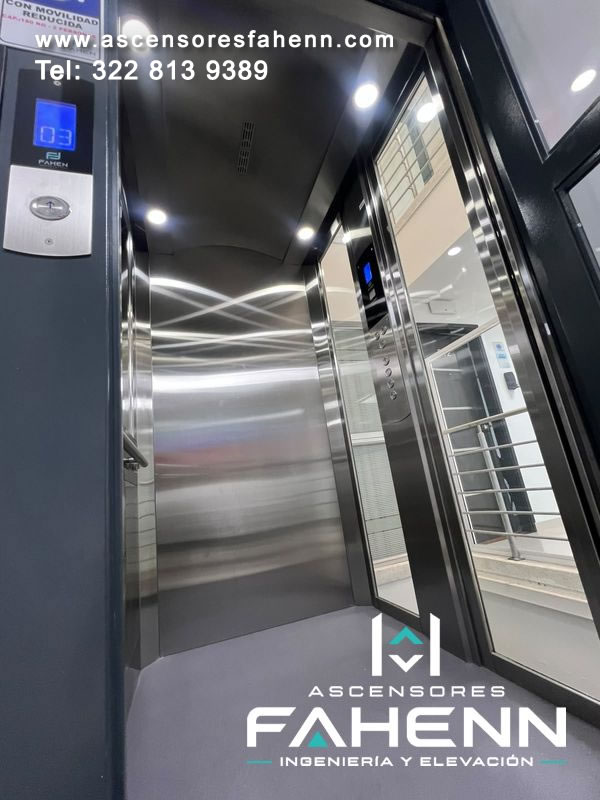 Ascensor accesible abierto con cabina panorámica de acero, vidrio lateral, luces superiores, botonera digital y pantalla exterior de piso 03.