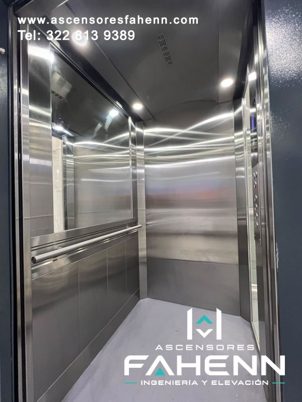Cabina de ascensor en acero inoxidable con espejo lateral, pasamanos doble, luces LED circulares, botonera derecha y piso gris continuo.