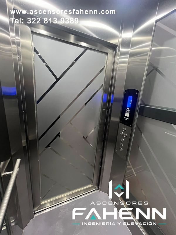 Cabina de ascensor con puerta de vidrio esmerilado y líneas geométricas, botonera digital de piso 3, espejo, pasamanos y paneles de acero.