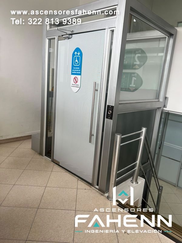 Elevador para personas con movilidad reducida con puerta de vidrio esmerilado, marco de aluminio, manija vertical y cerramiento lateral transparente.