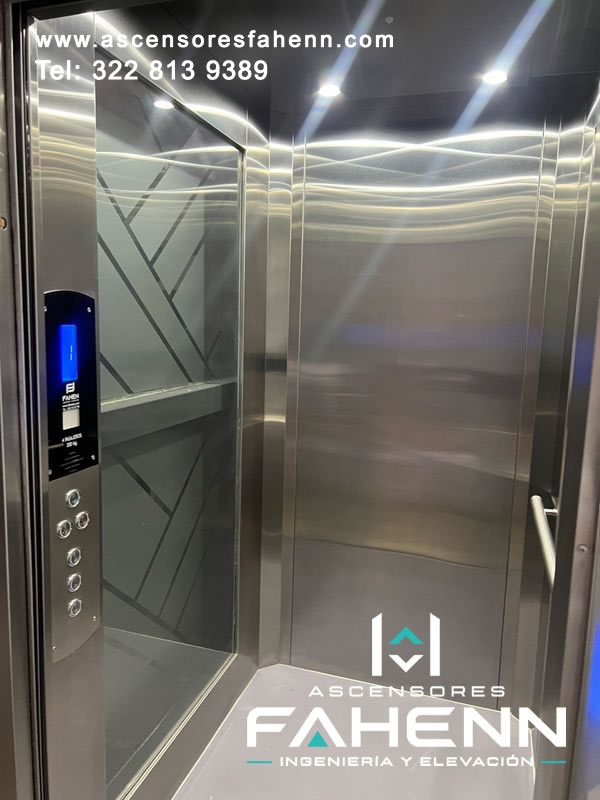 Cabina de ascensor con vidrio decorativo de líneas geométricas, paneles de acero, pantalla digital azul, pulsadores y pasamanos.