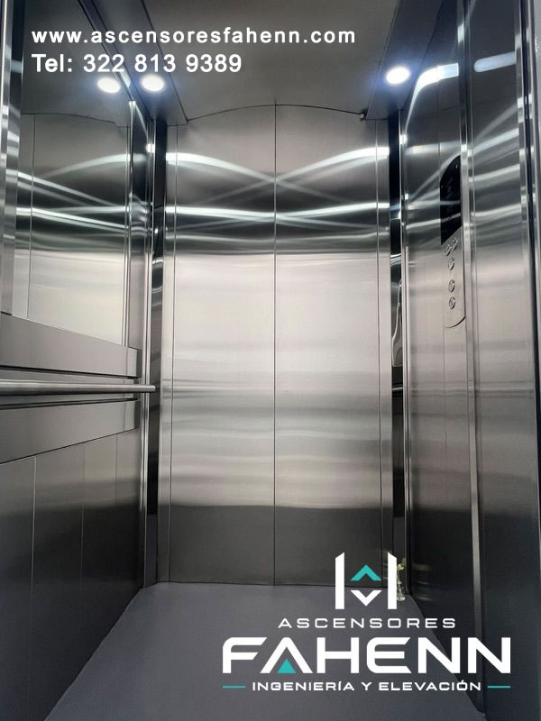 Cabina moderna de acero inoxidable con puertas automáticas cerradas, pasamanos doble, botonera lateral y luces circulares superiores.