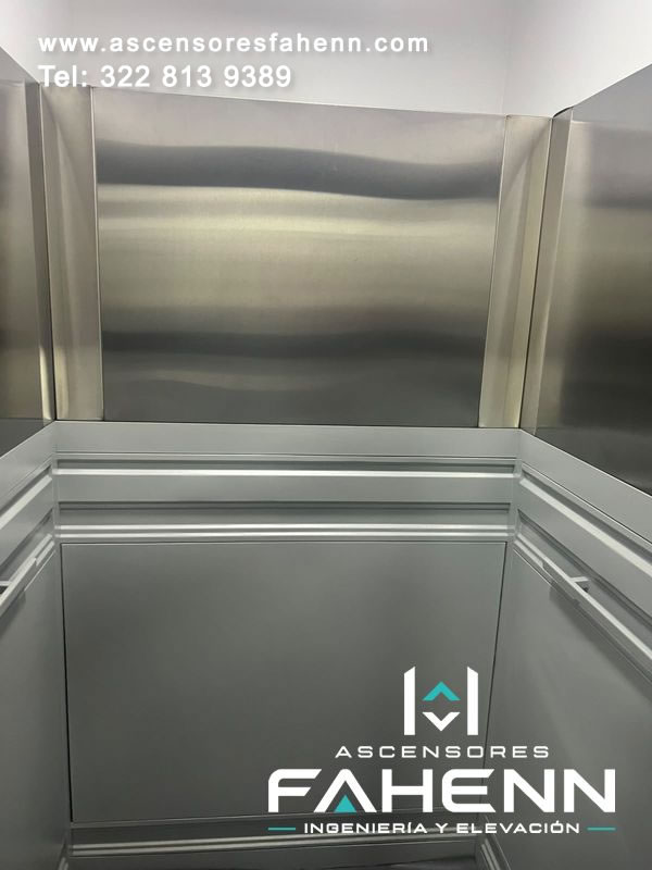 Cabina de ascensor con paneles inferiores grises, acero pulido superior, pasamanos doble alrededor y molduras metálicas horizontales.