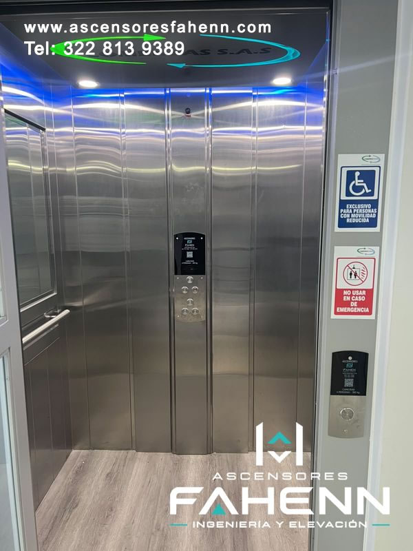 Cabina accesible de acero inoxidable con iluminación LED azul, panel de botones central, piso tipo madera y avisos de movilidad reducida.