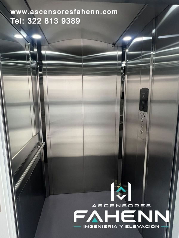 Cabina de ascensor en acero inoxidable con puertas centrales, espejo lateral, pasamanos horizontal, controles verticales y focos blancos superiores.