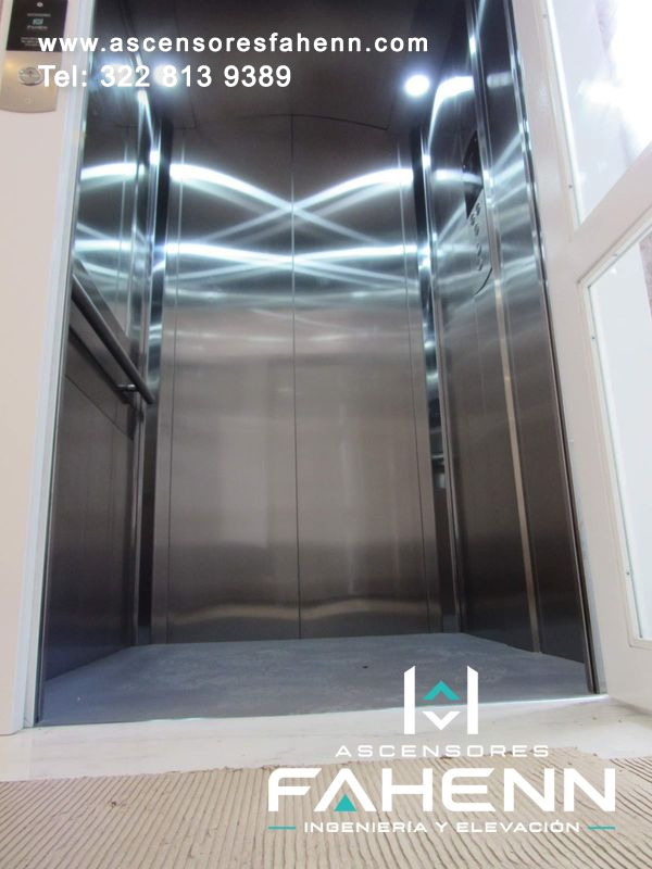 Cabina de ascensor en acero inoxidable con puertas centrales, pasamanos lateral, iluminación LED, botonera derecha y piso gris uniforme.