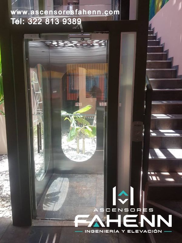 Ascensor para casa con cabina acristalada, perfiles negros, aberturas ovaladas, iluminación decorativa y escalera oscura contigua.
