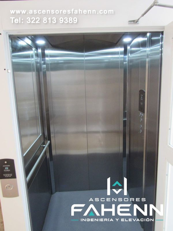 Cabina de ascensor en acero pulido con espejo izquierdo, pasamanos horizontal, panel de botones derecho, focos superiores y piso gris.