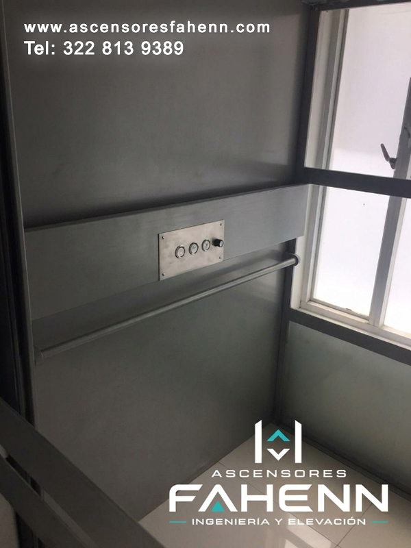 Cabina gris de ascensor residencial con ventana lateral amplia, pasamanos metálico, cuatro controles alineados y revestimientos sobrios.