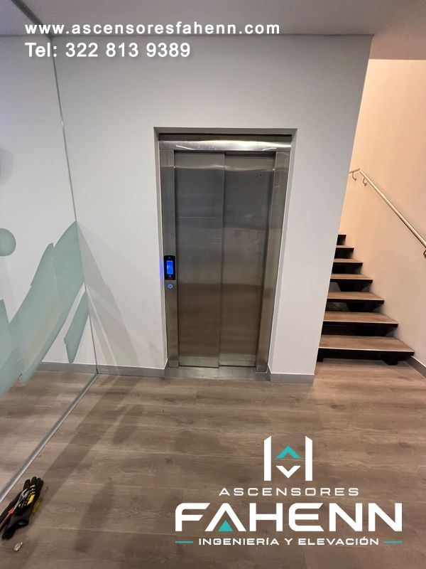 Puertas automáticas de ascensor en acero instaladas junto a escalera, con indicador azul, piso tipo madera y cerramiento lateral de vidrio.